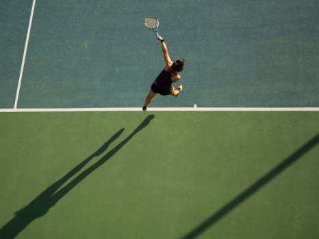 A high-angle, aerial view of a female tennis player in mid-serve. She is captured mid-jump with her racket arm extended high above her head.