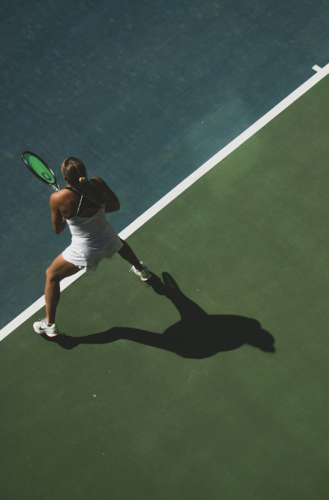 A high-angle photograph shows a female tennis player in a white outfit and visor, holding a racket, on a green tennis court. She is in a ready stance near the white baseline, with her long shadow cast across the green surface.